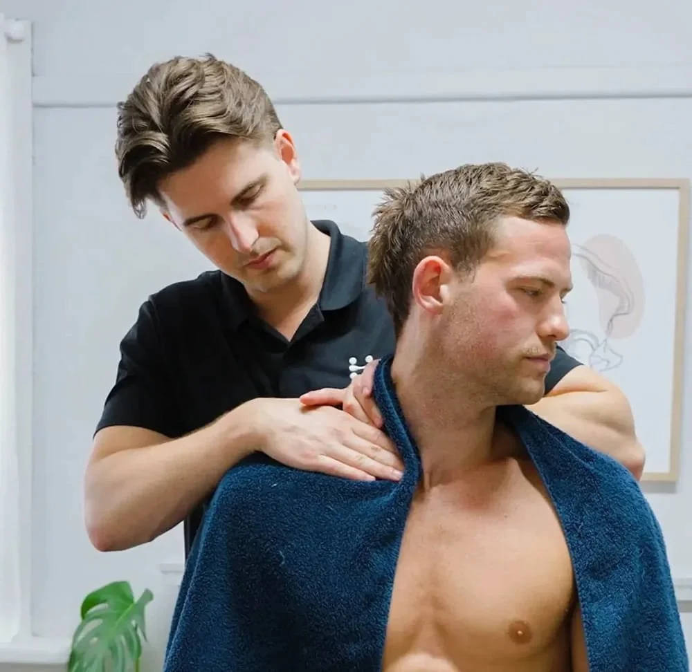 Therapist performing a shoulder and neck massage on a seated man draped with a towel in a clinic setting.