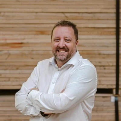 A man with a beard and short hair smiling in front of a wooden background, wearing a white dress shirt with folded arms.