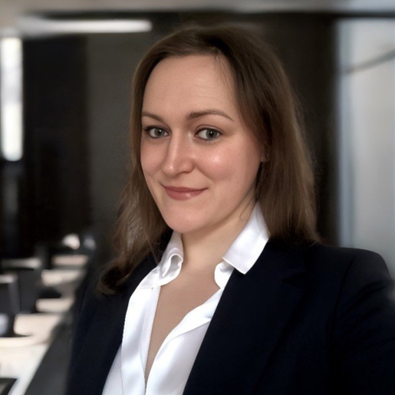 A young woman with long brown hair, wearing a white collared shirt and a black blazer, smiling at the camera in a professional setting.