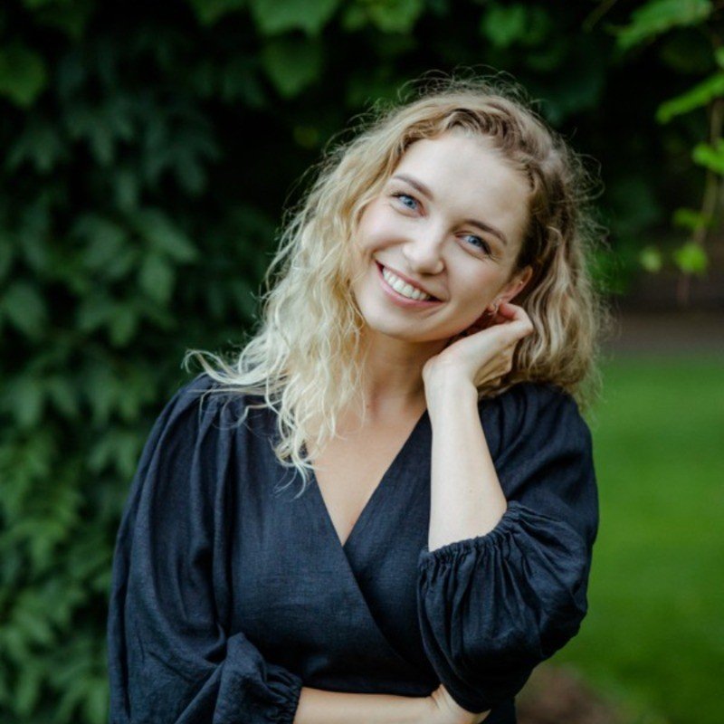 A young woman with curly blond hair smiling outdoors, dressed in a dark top, with green foliage in the background.