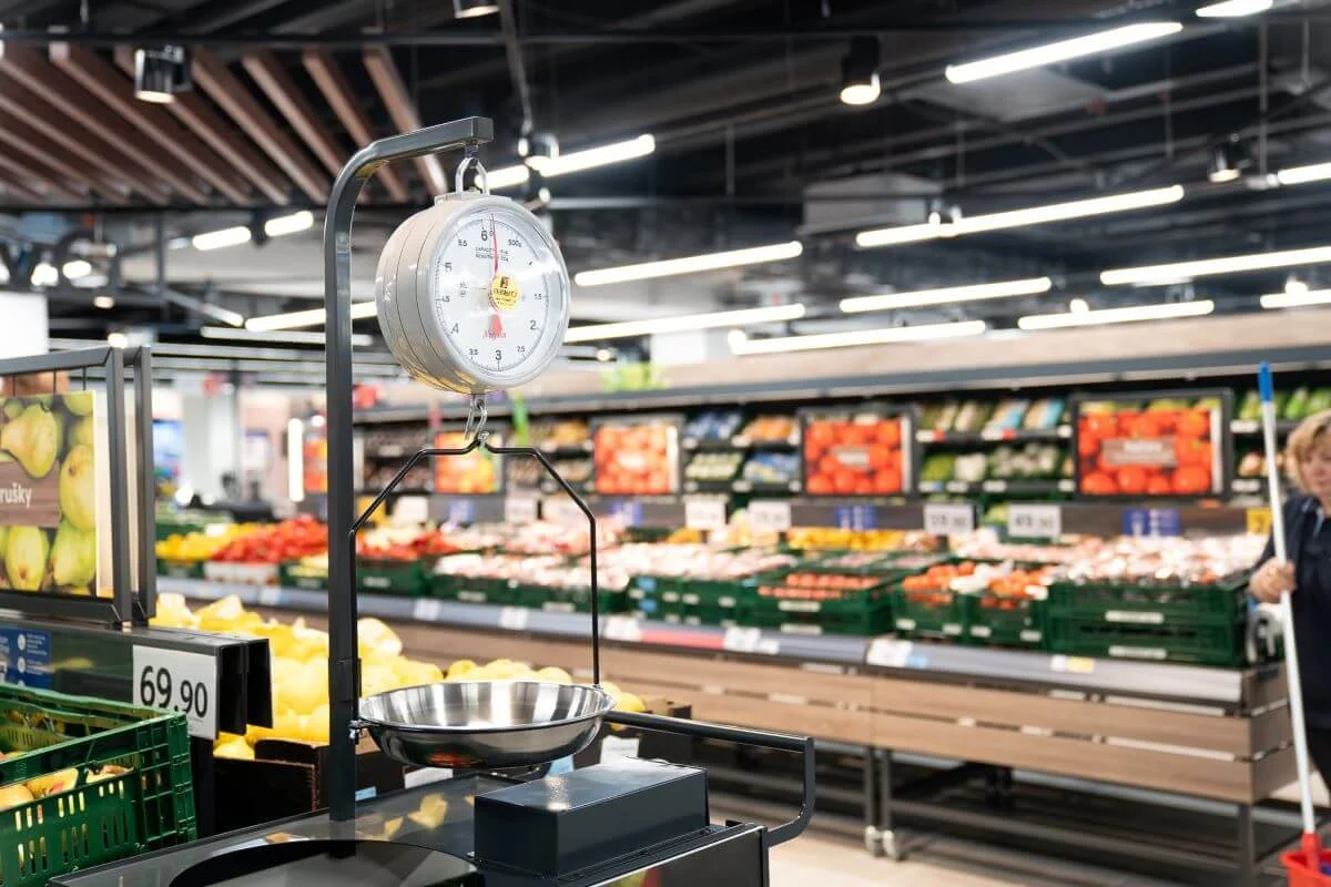 A supermarket produce section with a hanging scale and basket, surrounded by displayed fruits and vegetables.