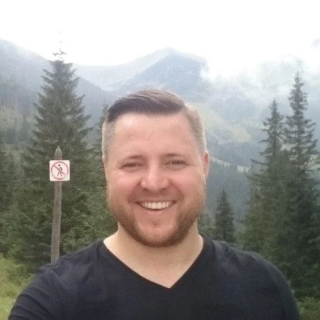 A smiling man with short hair and a beard outdoors, with trees and mountains in the background.
