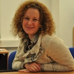 A woman with curly hair smiling, sitting at a table in an indoor setting.