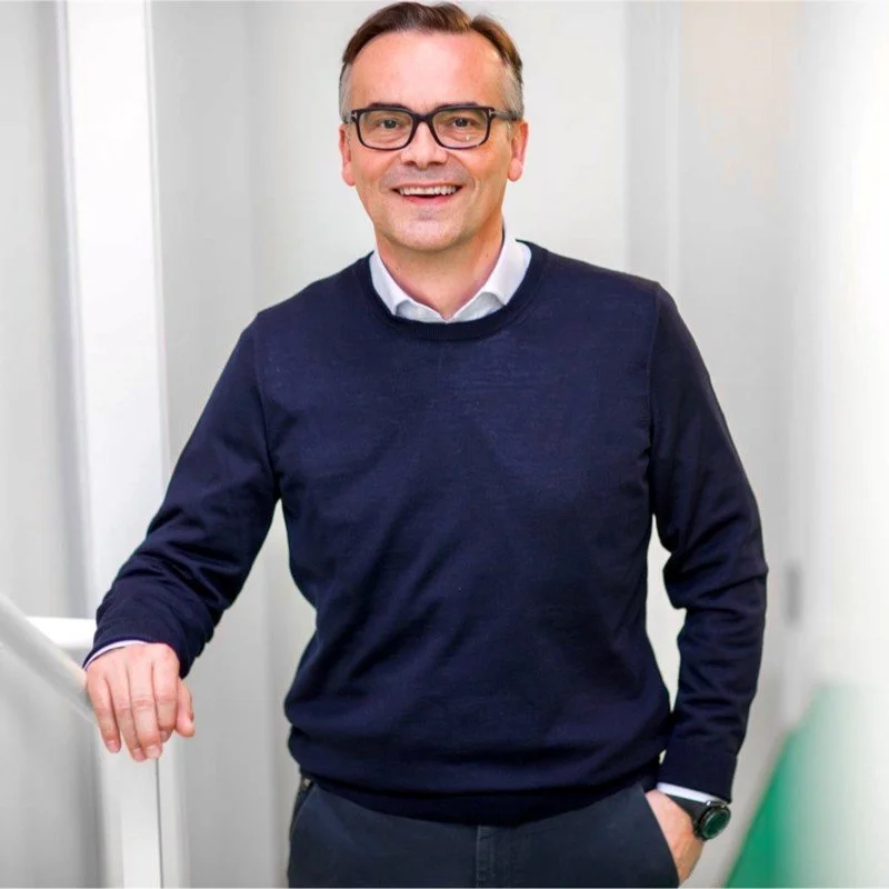 A man wearing glasses, a navy sweater, and dark pants smiling indoors, standing near a desk or counter.