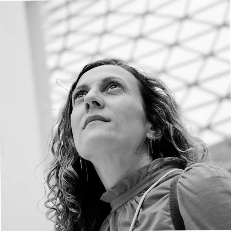 A woman with curly hair looking upward indoors with a geometric ceiling design in the background.