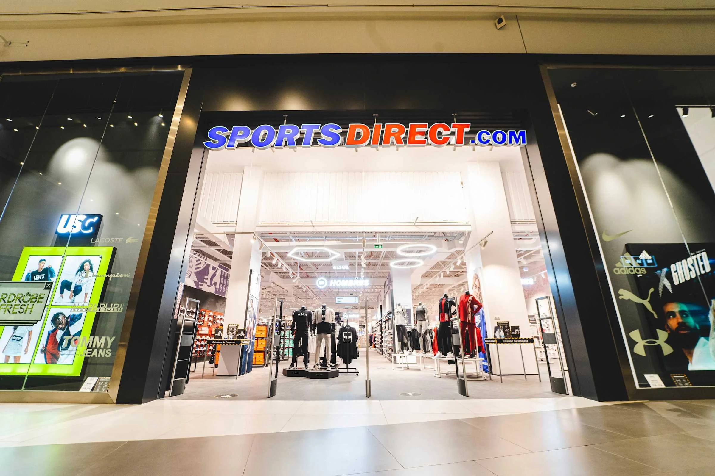 Entrance to a Sports Direct retail store with mannequins dressed in sportswear and athletics equipment visible inside.