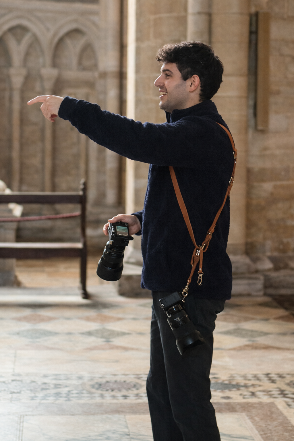A young man in a black fleece jacket standing indoors with a DSLR camera hanging from his neck and a camera in his right hand, pointing with his left hand in a pointing gesture.