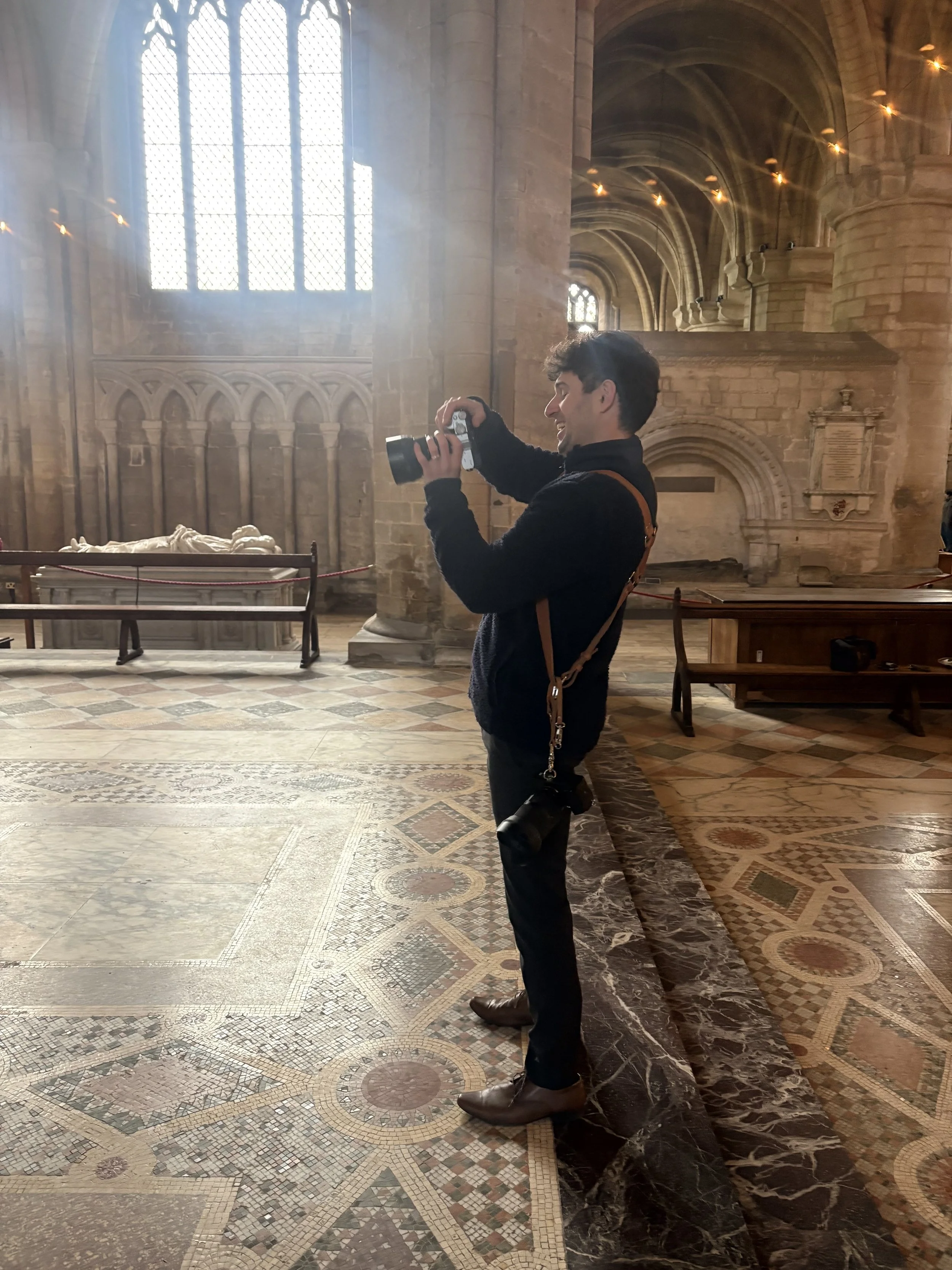 A young man with dark hair holding a camera inside a historic stone church or cathedral, with stained glass windows and intricate tile flooring.
