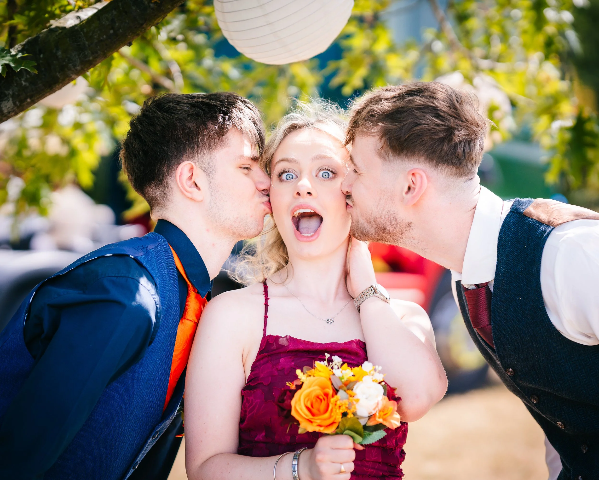 Woman holding a bouquet while two men kiss her on the cheek at a wedding