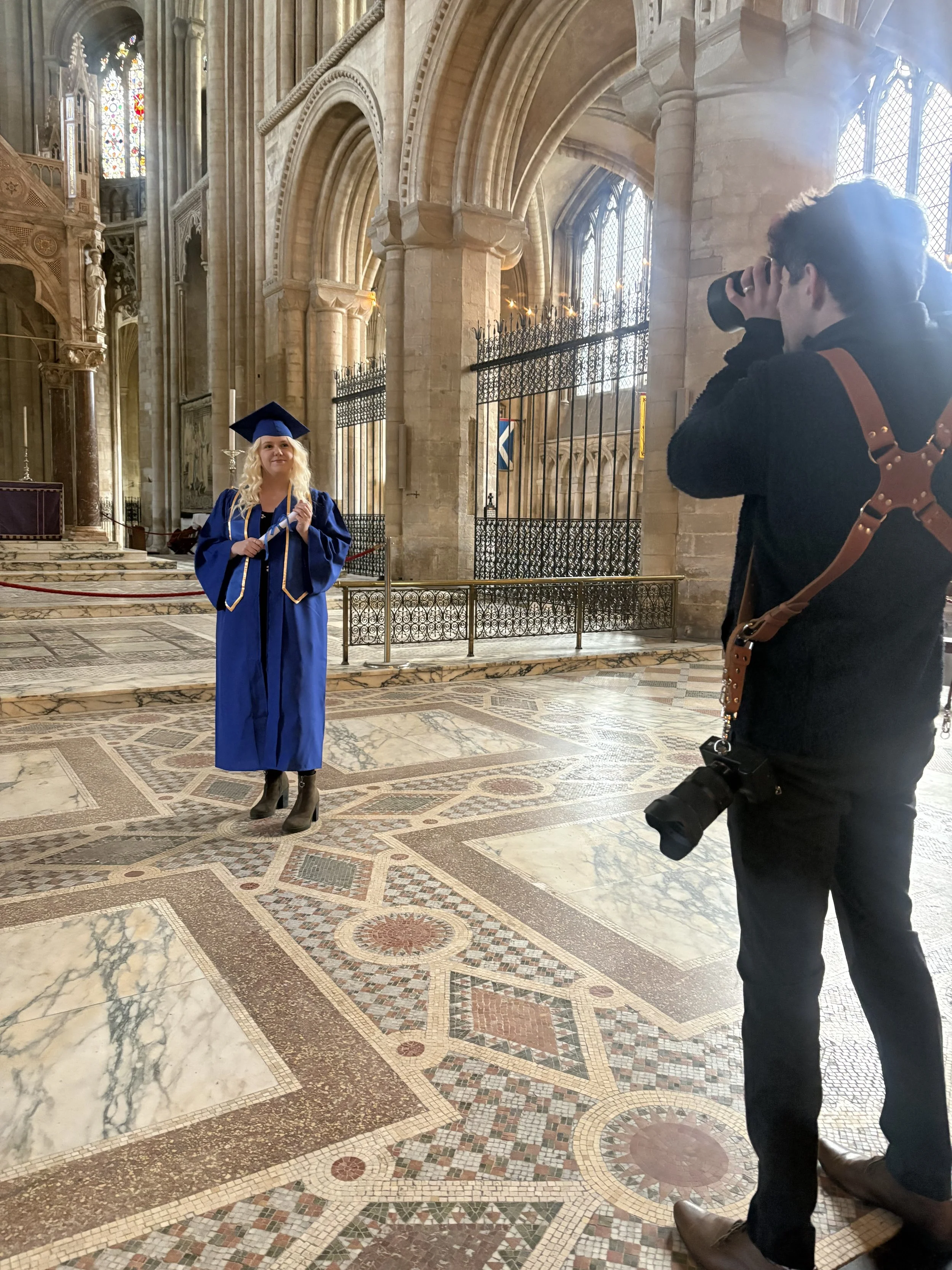A woman in a blue graduation gown and cap stands inside a grand cathedral, posing for a photo. A photographer is taking her picture from the right side of the image.