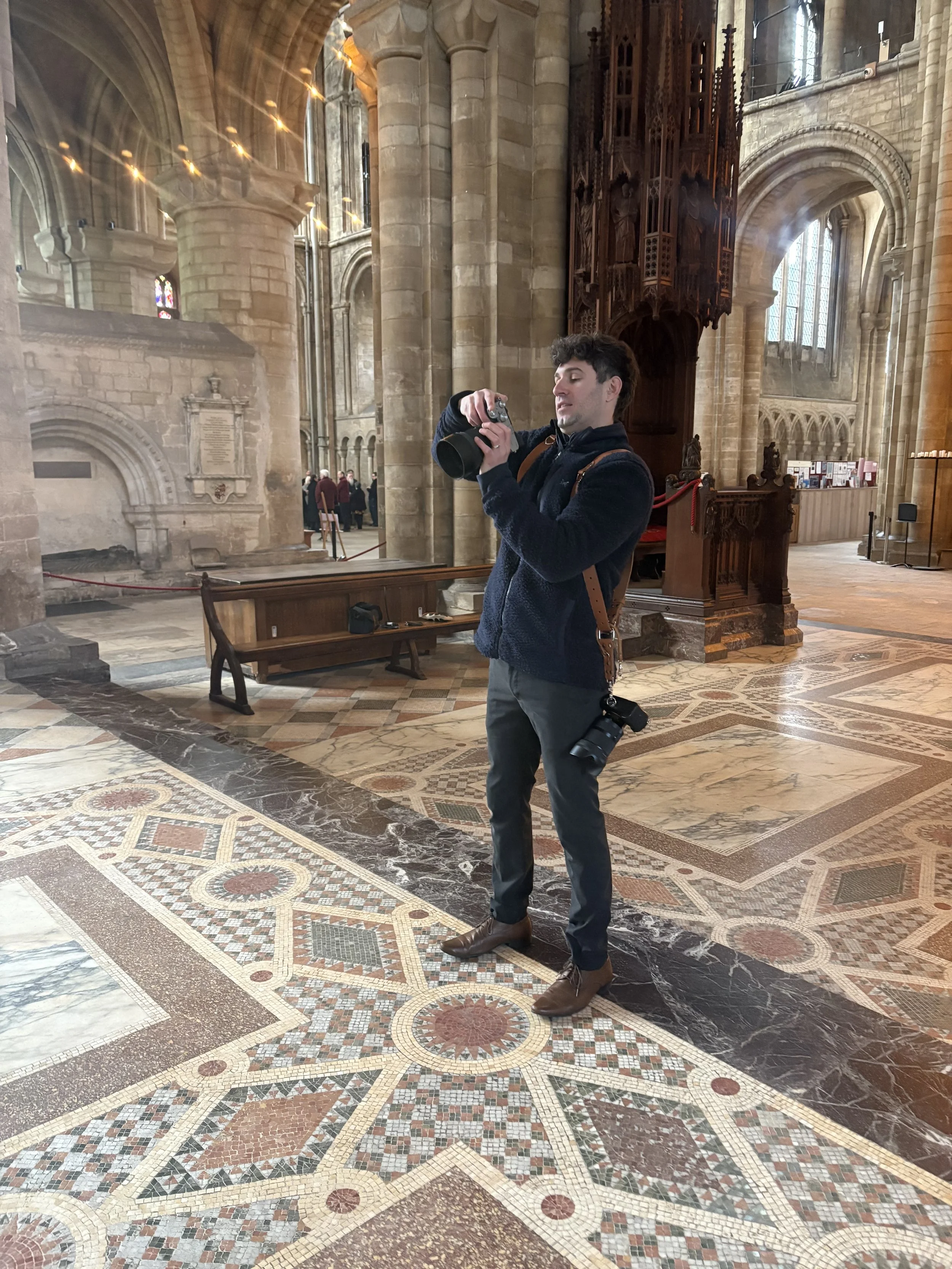 A man taking a photo inside a historic church or cathedral with ornate, colorful tile flooring and stained glass windows.
