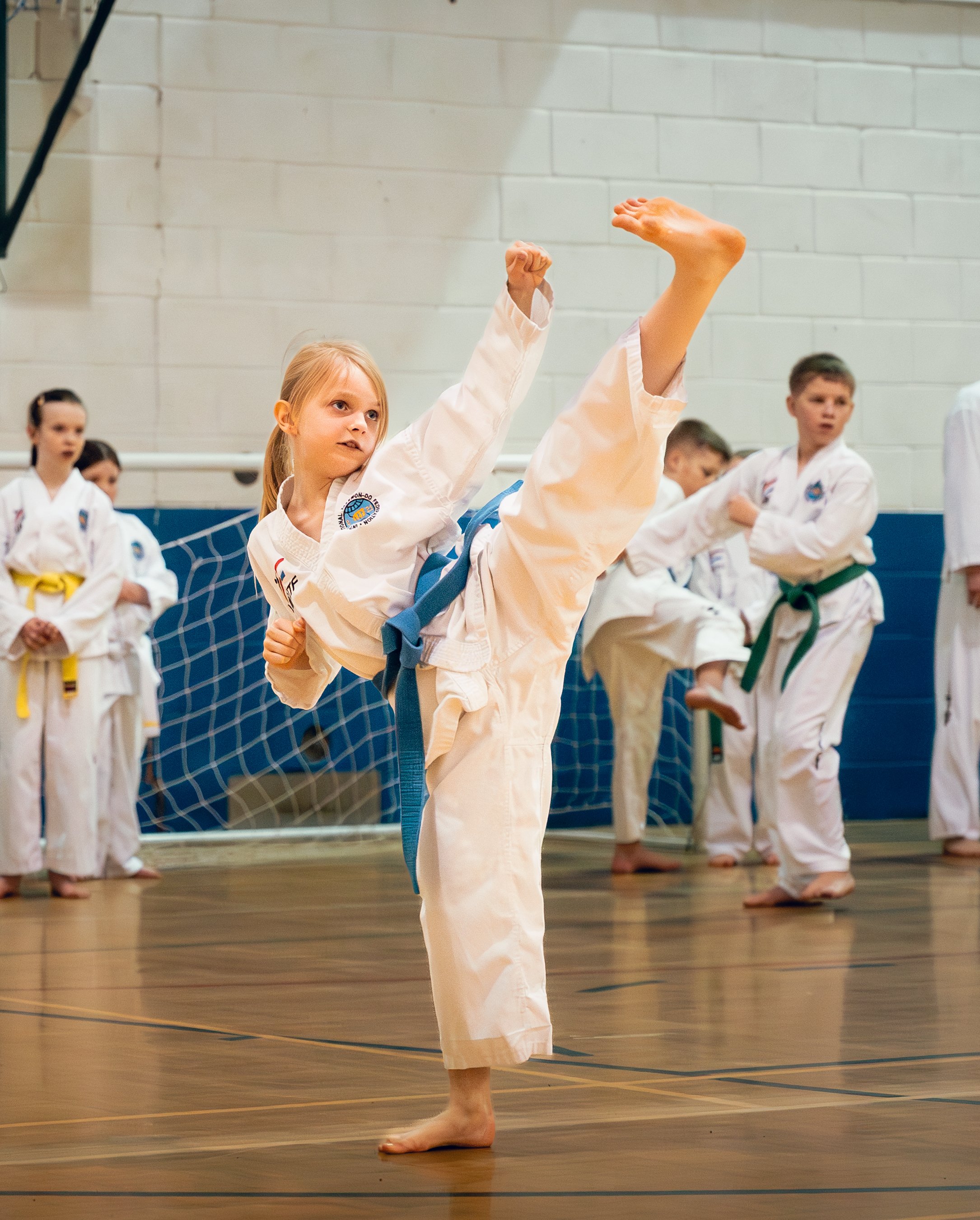 Young girl performing a high kick during a karate class with other children in the background.