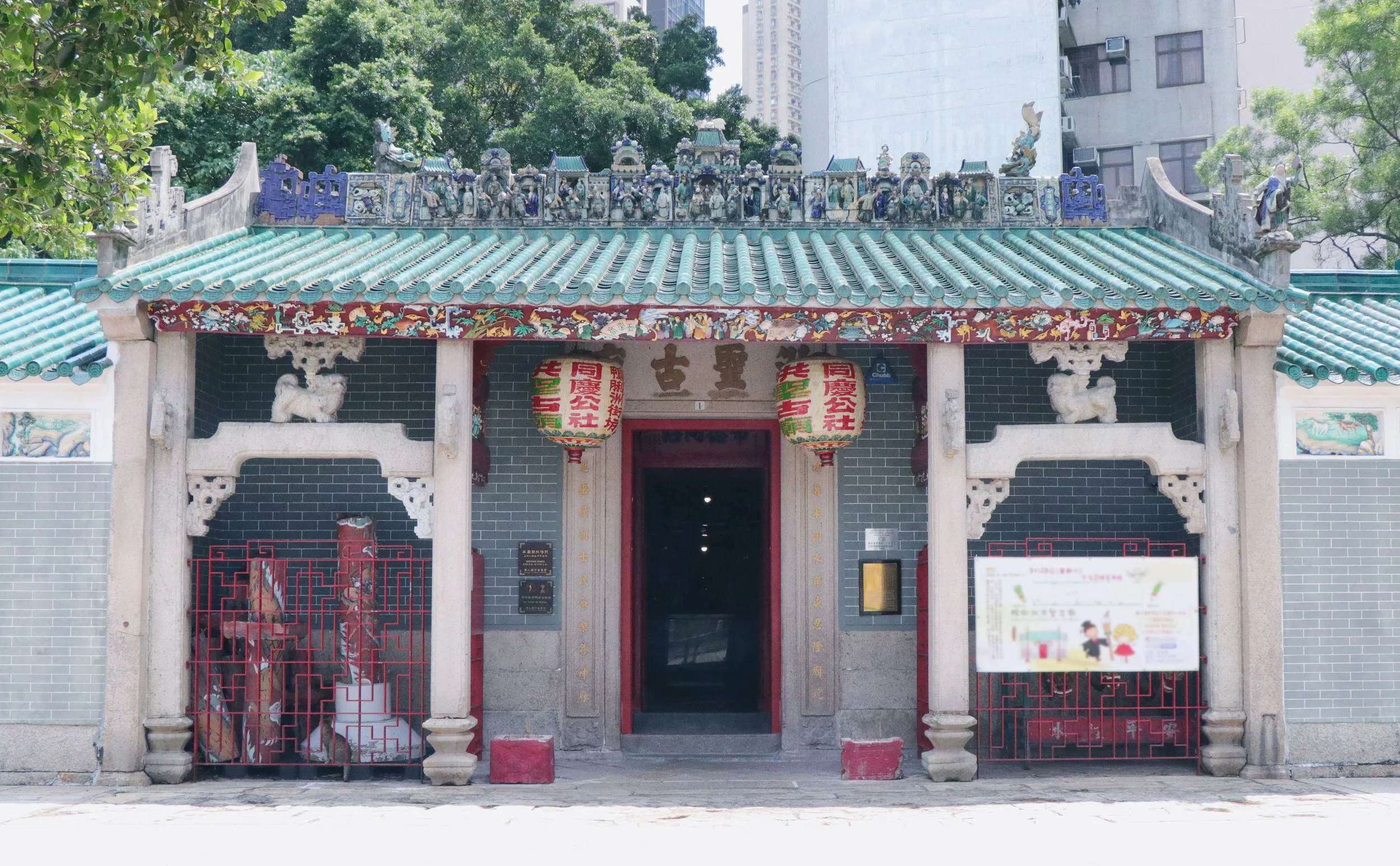 Front view of a traditional Chinese temple entrance with a decorative tiled roof, ornate carvings, hanging lanterns, and a gate. Tall buildings and green trees are in the background.