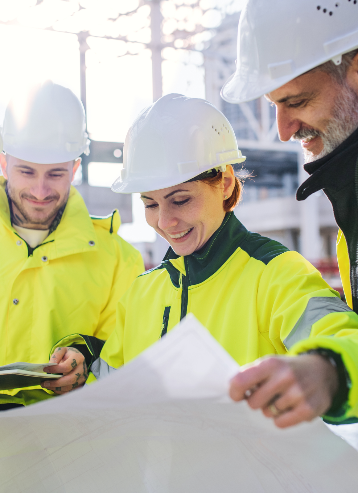 Construction workers in yellow safety jackets and white helmets reviewing plans at a construction site.