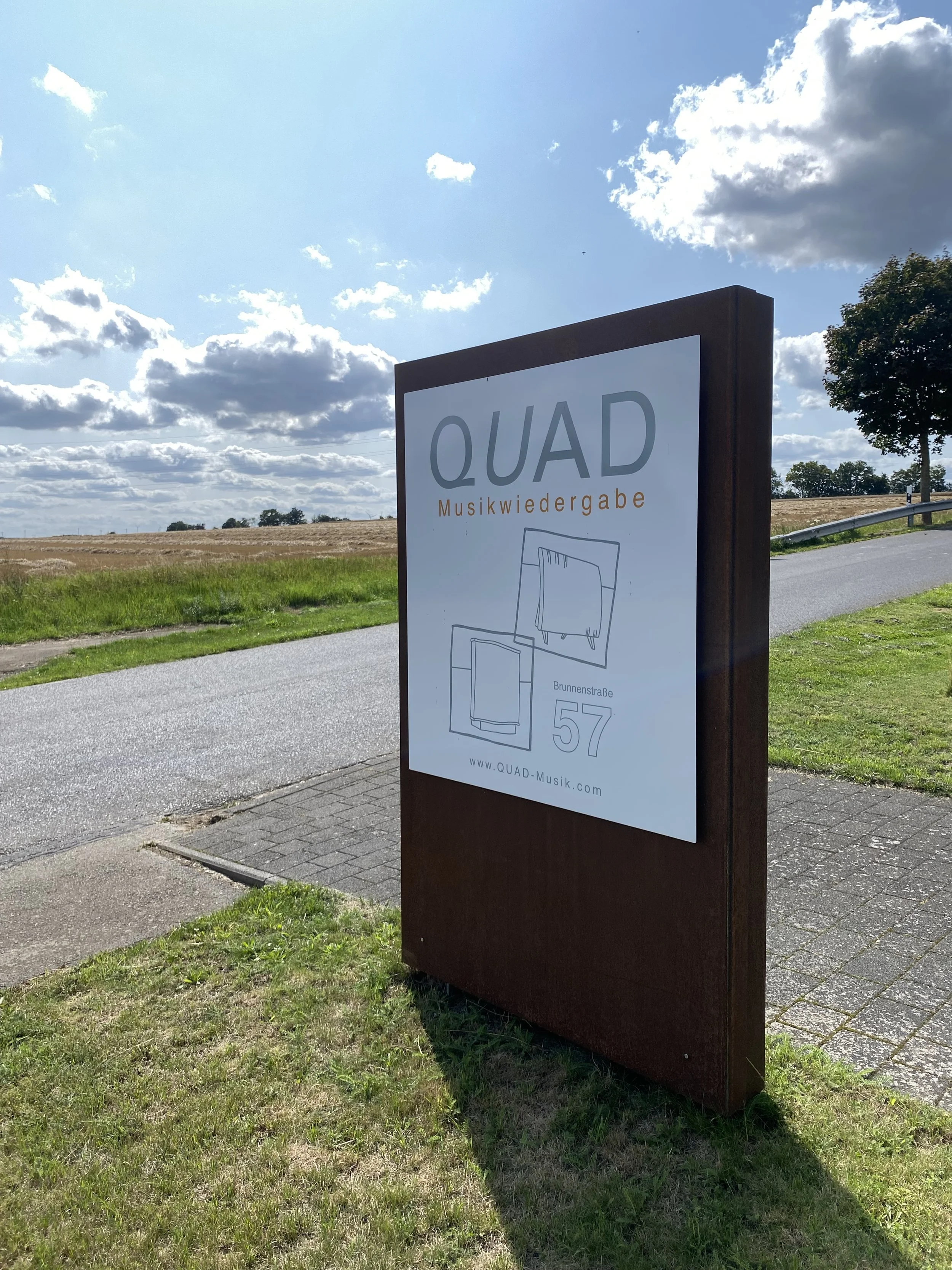 Hinweisschild für eine Musikvergabe namens QUAD an der Straße mit Feldern im Hintergrund, der das Logo, die Adresse und die Webseite zeigt.