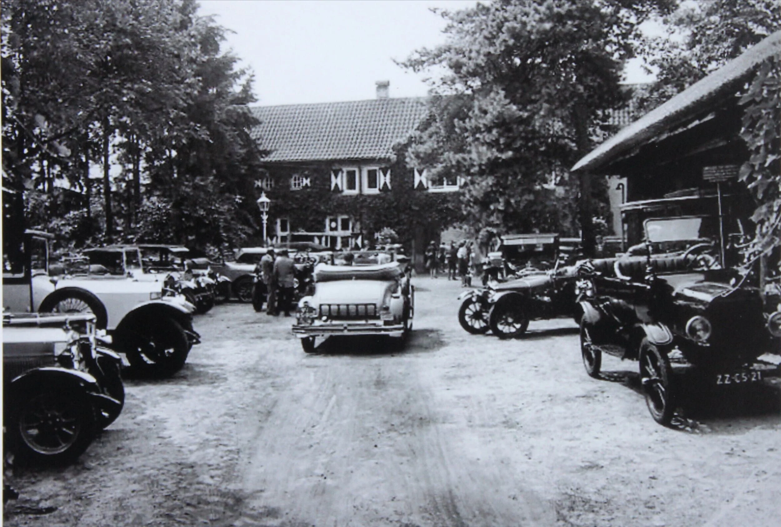 Black and white photo of a vintage car show or gathering in a parking area with several classic cars and motorcycles. People are walking around and looking at the vehicles, with trees and a building in the background.