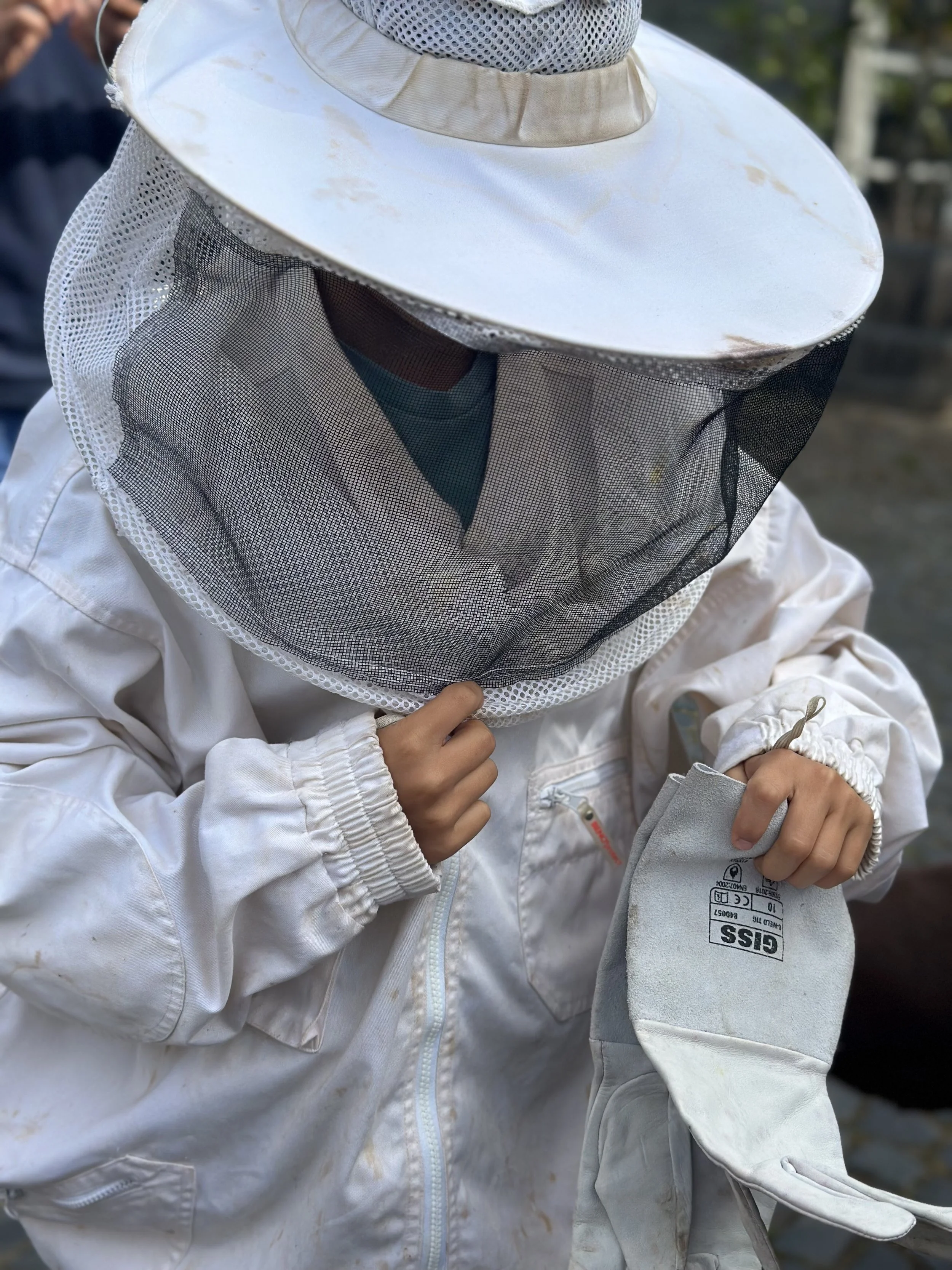 Child wearing a white protective beekeeping suit and hat with a black mesh veil, holding a worn white beekeeping glove.