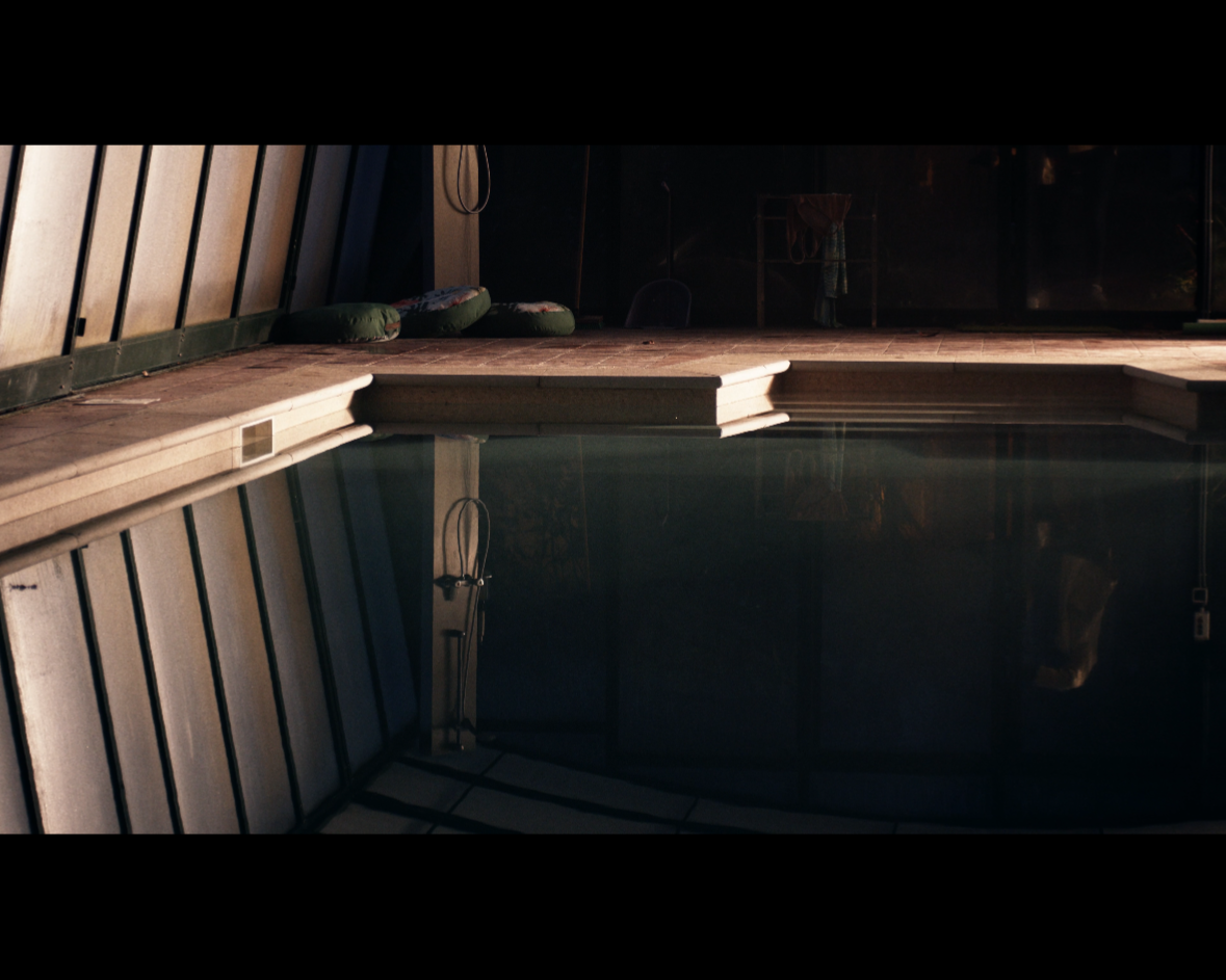 Indoor swimming pool with dark water, surrounded by a wooden deck and a wall with diagonal wooden panels; green cushions are placed on the deck.