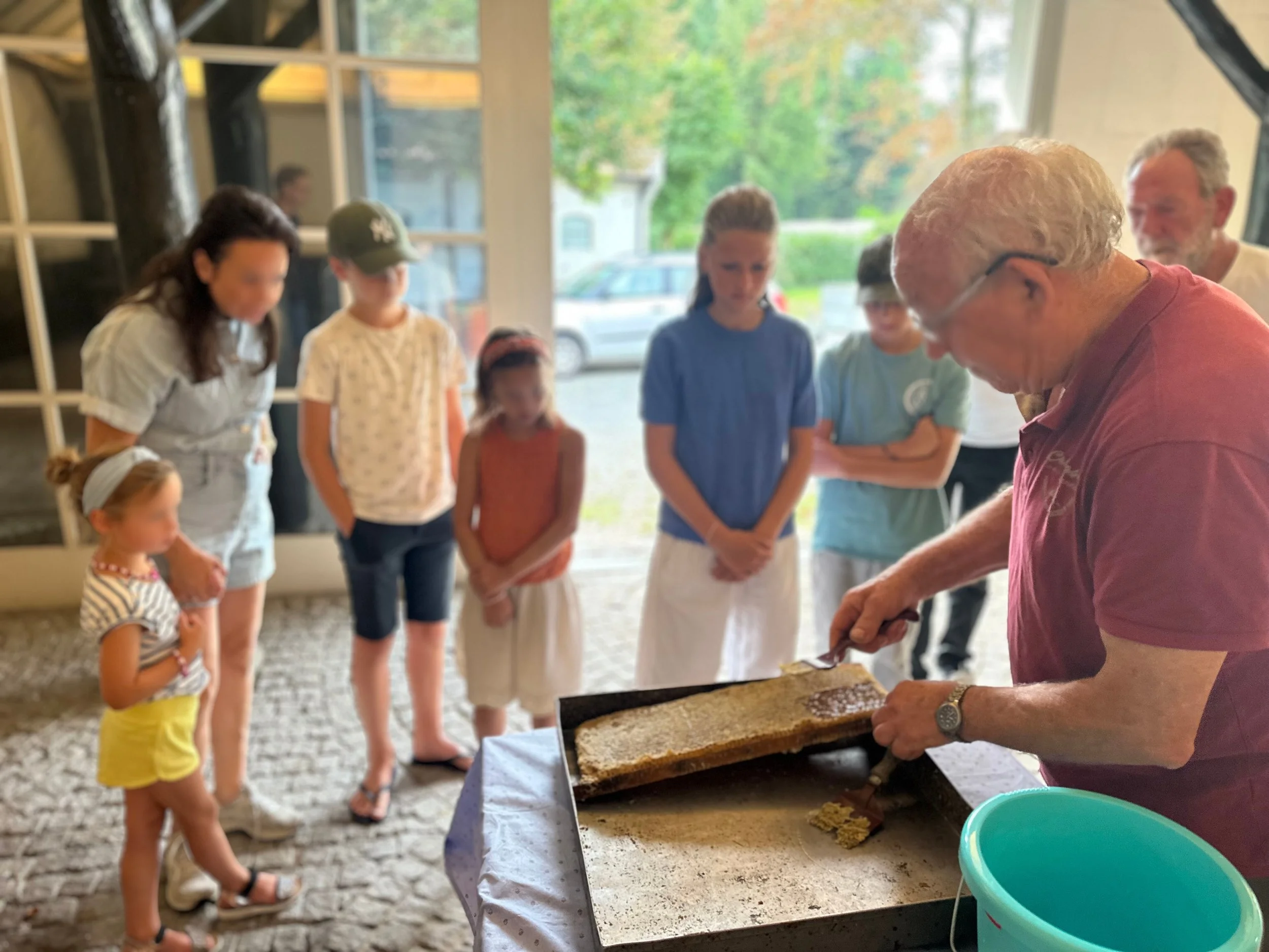 A man demonstrating honey harvesting to a group of children and adults inside a shop or cafe, with the man using a slicing tool on a honeycomb frame.