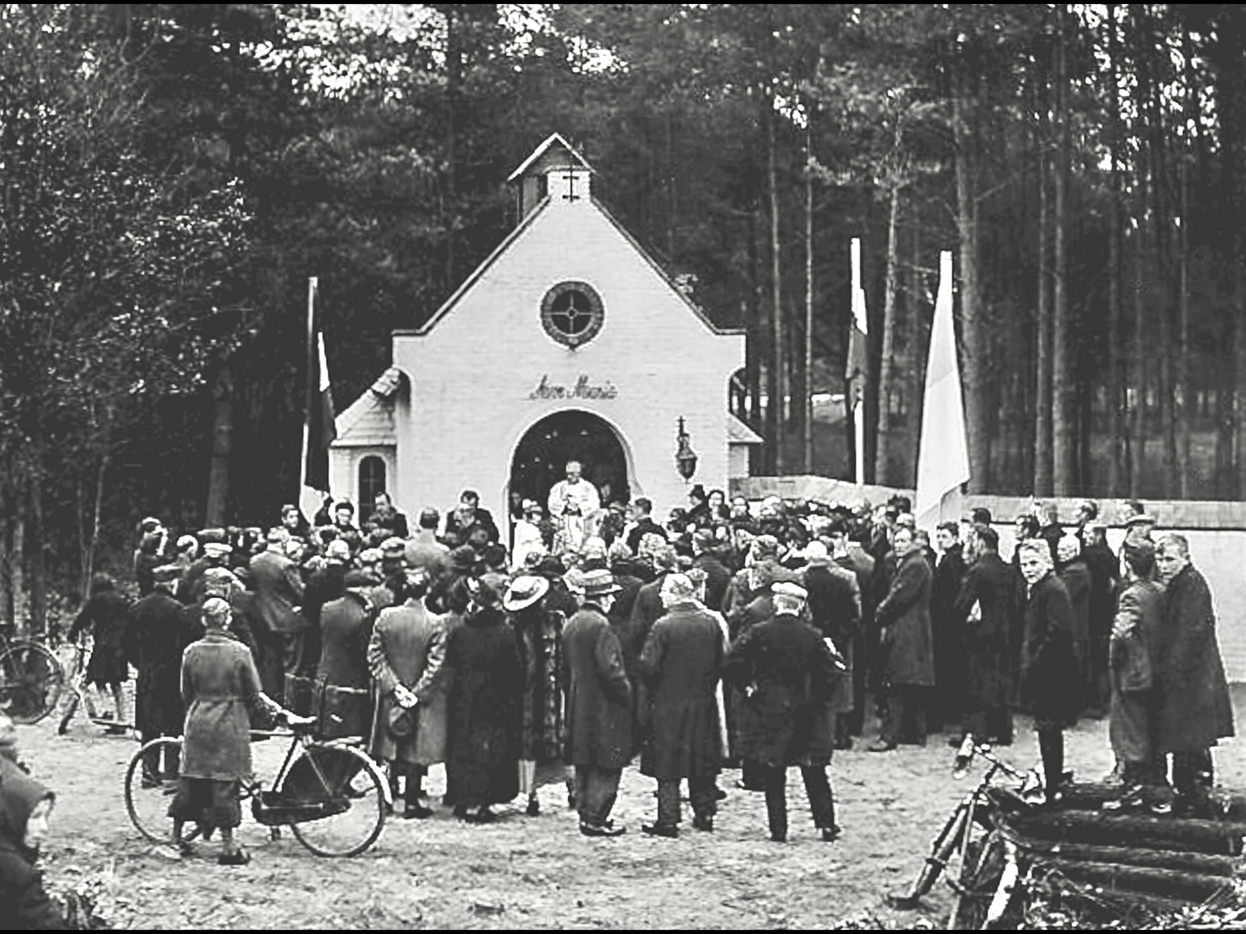 A black and white historical photograph of a gathering of people in front of a small church building with a cross on top. The crowd is standing outdoors, many wearing coats and hats, with some bicycles visible in the foreground. The church has the words 'The House' written above the entrance.