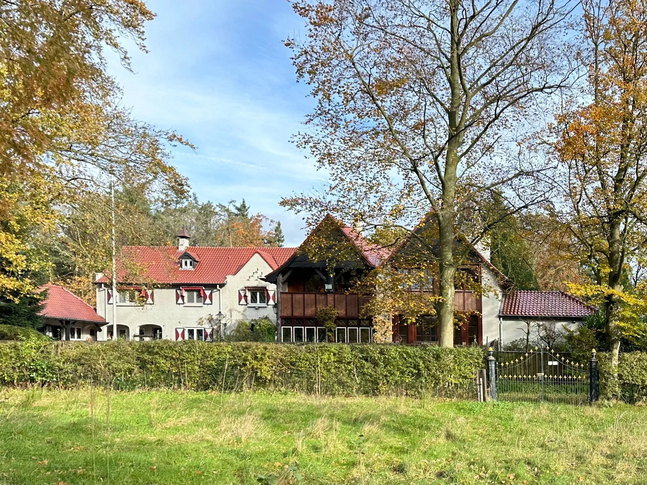 A large house with white walls, red-tiled roof, and multiple windows, surrounded by trees with autumn foliage and a green lawn in the foreground.