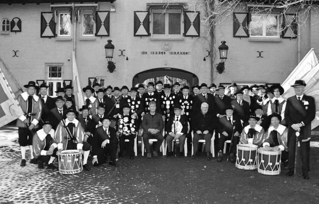A large group of people, mostly men, dressed in traditional European costumes, gathered outside a building with shuttered windows and lanterns, some seated and others standing, with musical instruments such as drums in the front.