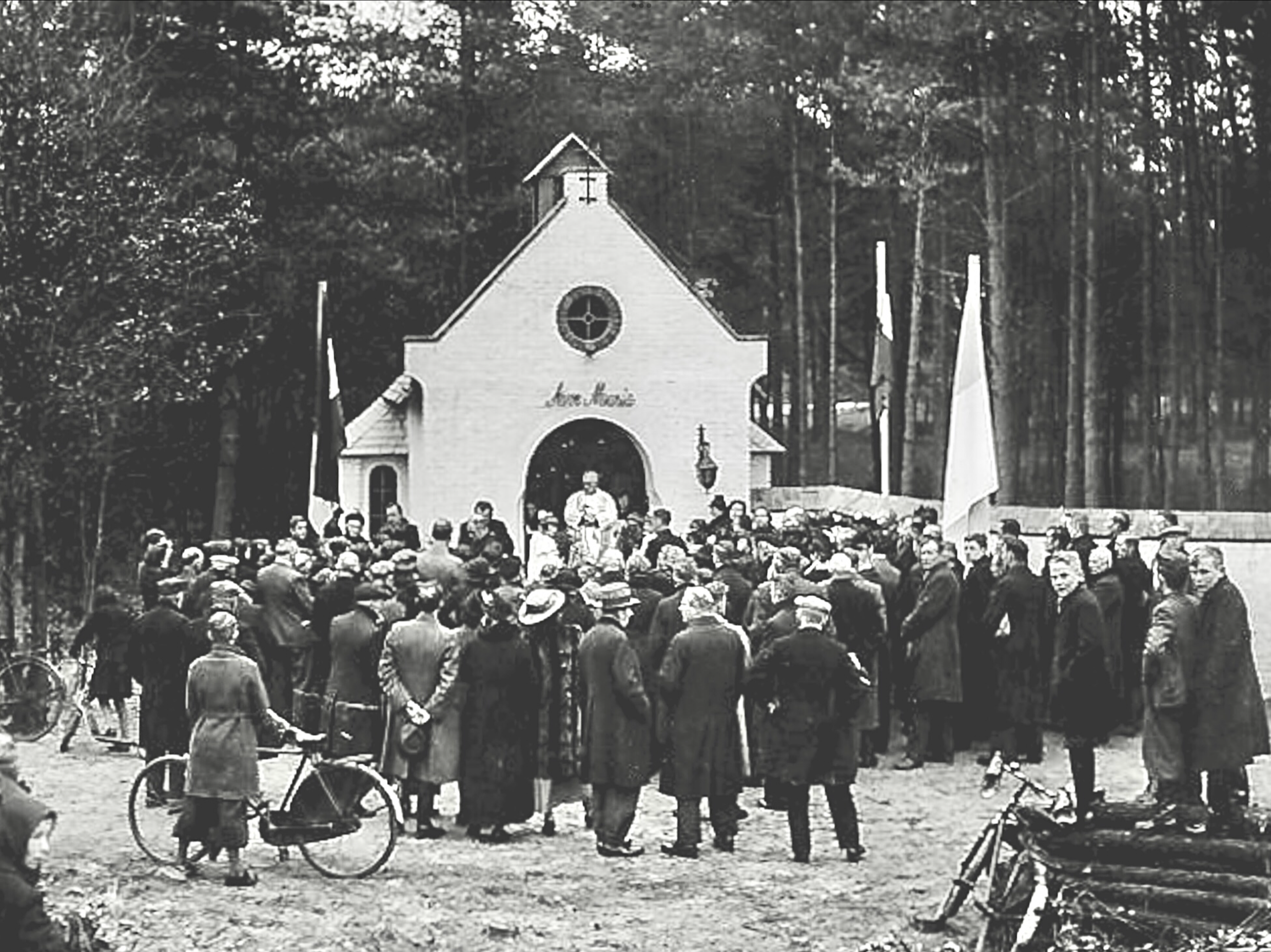 A large gathering of people in front of a small chapel-like building outdoors, with trees in the background. Several flags are visible, and some individuals are on bicycles. The crowd appears to be attending an event or ceremony.
