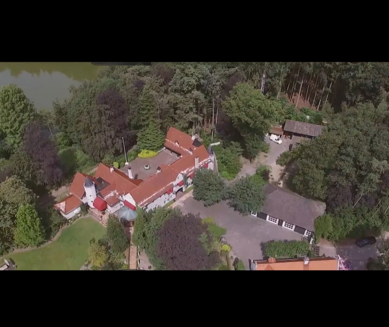 Aerial view of a large mansion with red roofs, surrounded by green trees, with a pond on the left, and a gravel driveway leading to outbuildings and parked cars.