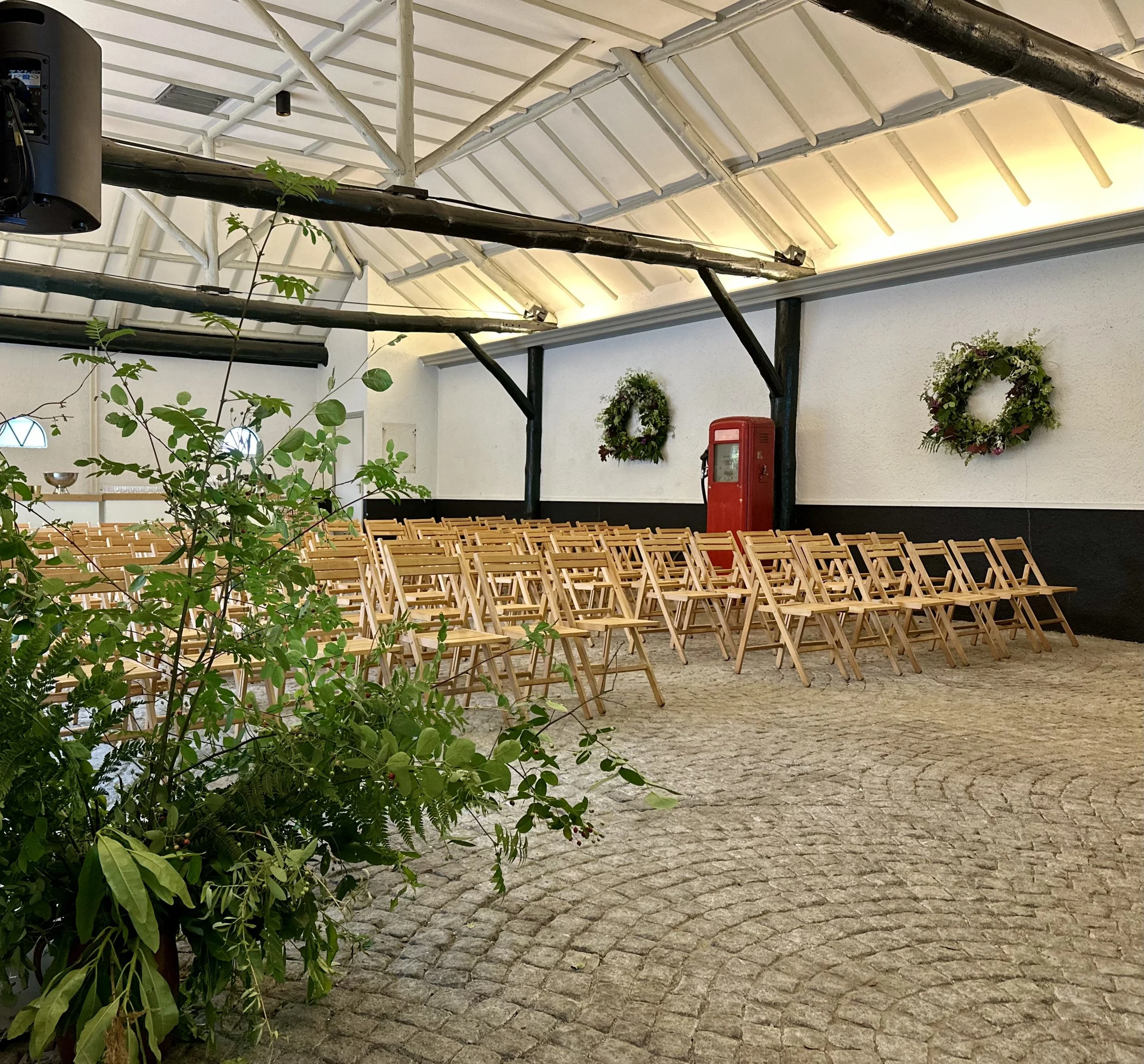 An indoor event space with wooden folding chairs arranged in rows, decorated with wreaths on the wall, and a vintage red petrol pump. The space has a vaulted ceiling with exposed beams and a stone-tiled floor. Green plants are in the foreground.
