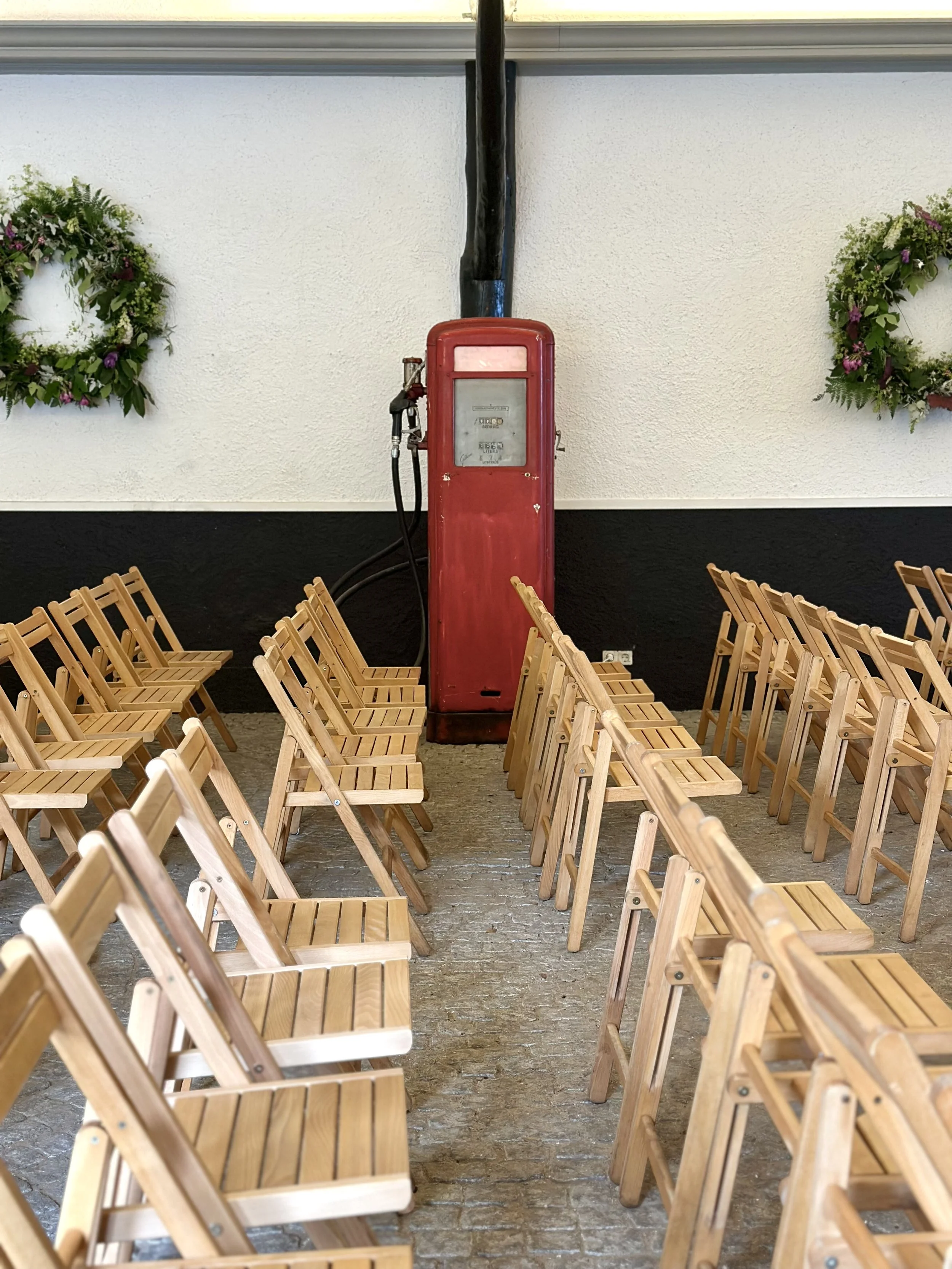 Empty wooden chairs arranged in rows facing a vintage red gas pump against a light-colored wall decorated with green plants.