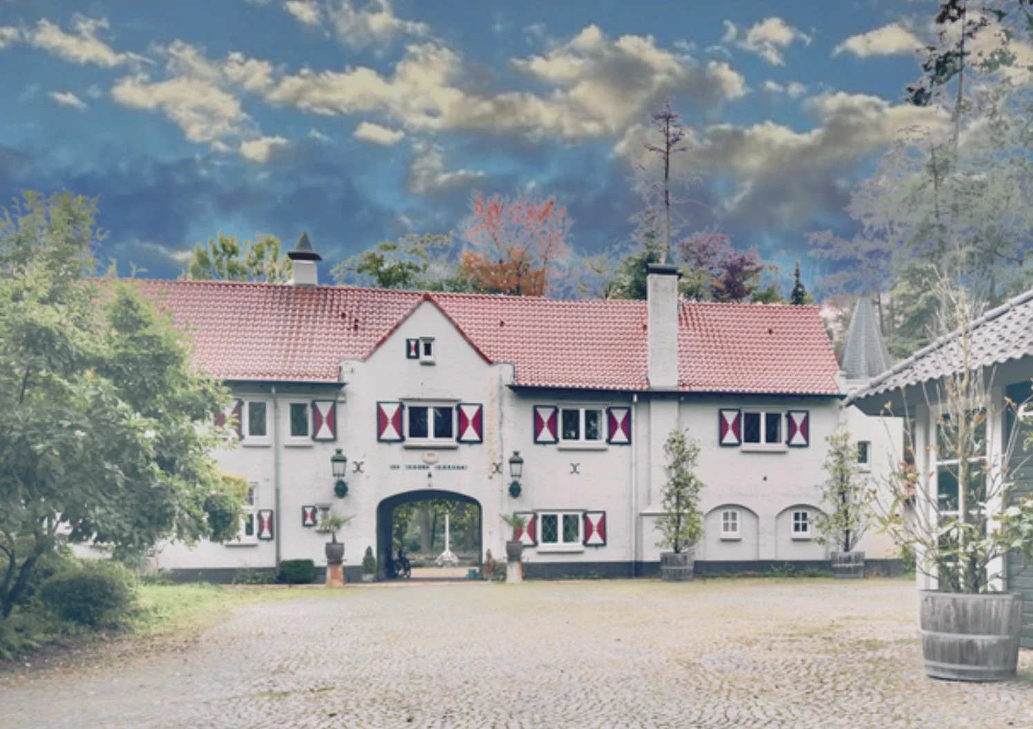 A white house with red shutters and a red tile roof, an arched driveway in the center, and cobblestone pavement in the foreground. There are trees and a partly cloudy sky in the background.