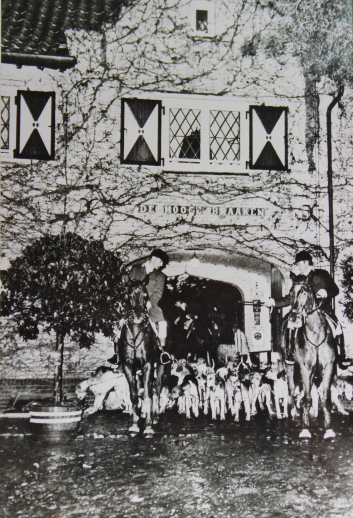 Black and white historic photo of a group of dogs and two individuals on horseback in front of a building with a sign that reads 'DE 1001 BREKEN.'