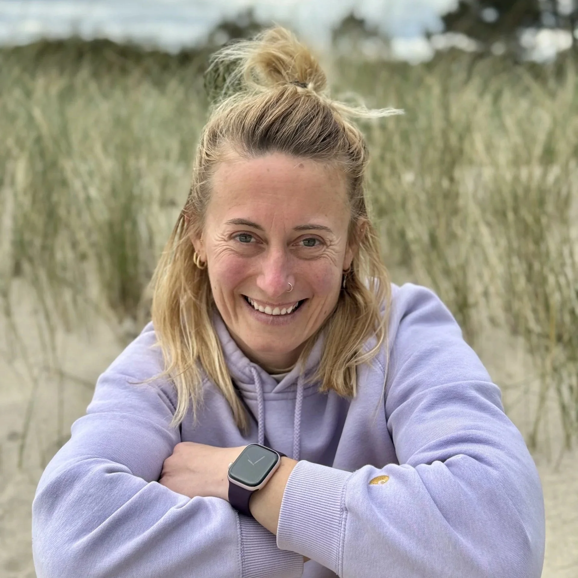 Une femme souriante avec des cheveux blonds attachés en haut, portant un sweat à capuche violet et une montre connectée au poignet gauche, posée sur une plage de dunes de sable avec de l'herbe.