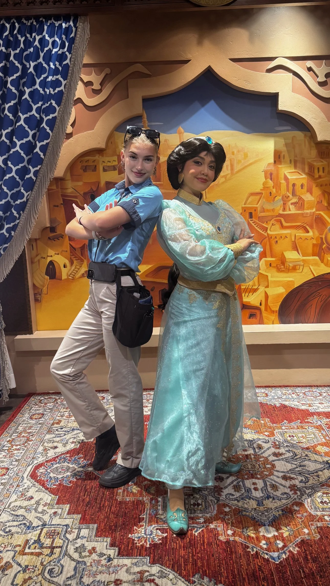 A woman dressed as Princess Jasmine from Aladdin standing in front of a painted desert backdrop, and a young woman in a blue uniform with sunglasses on her head standing next to her, both posing with arms crossed.
