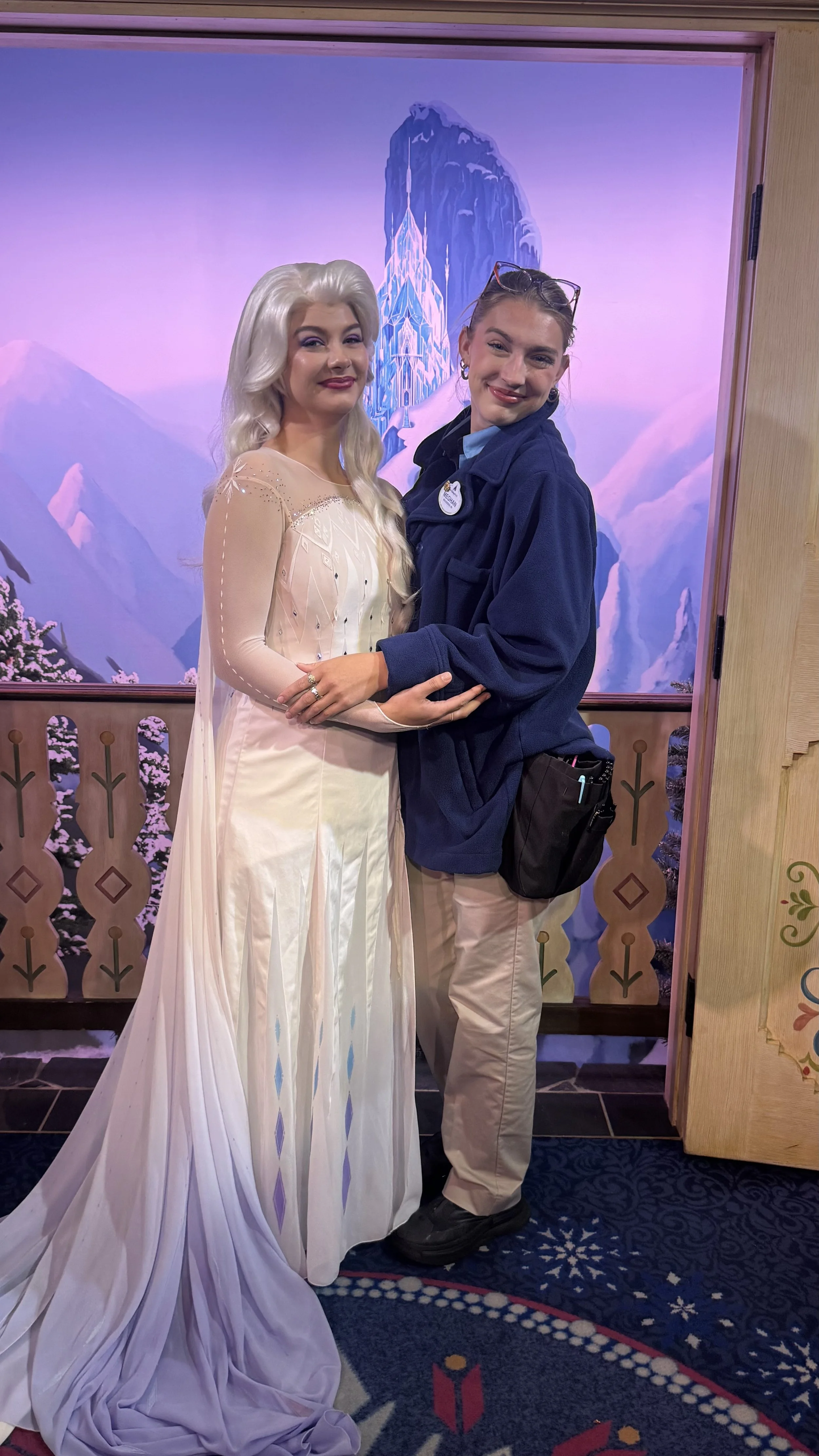 Two women standing close together, one dressed as Elsa from Frozen with a white gown and platinum blonde hair, and the other in a blue jacket with glasses on her head, in front of a fantasy mountain background with snow and ice.