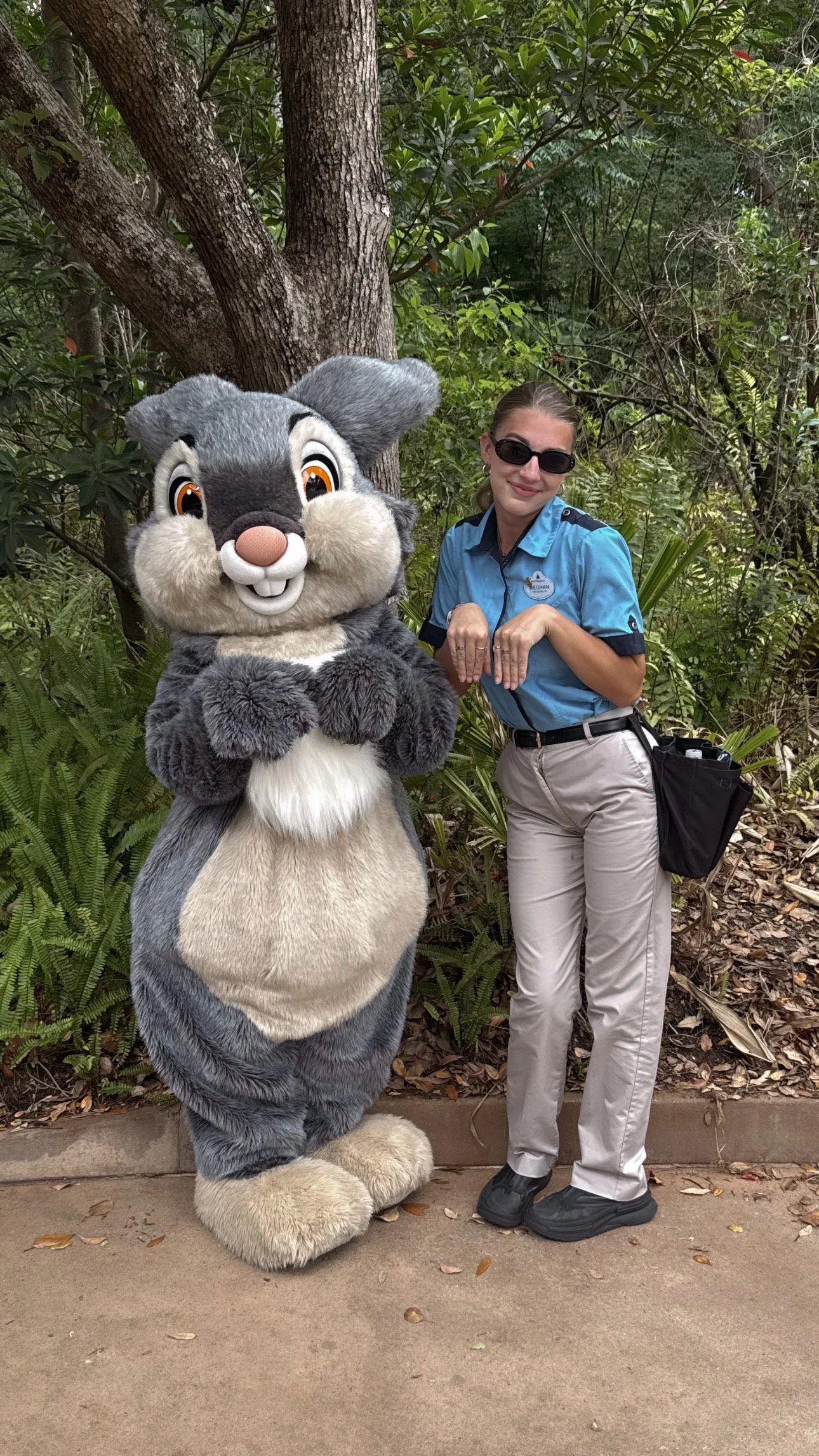A person wearing sunglasses and a blue shirt standing next to a person in a rabbit costume in front of green foliage.