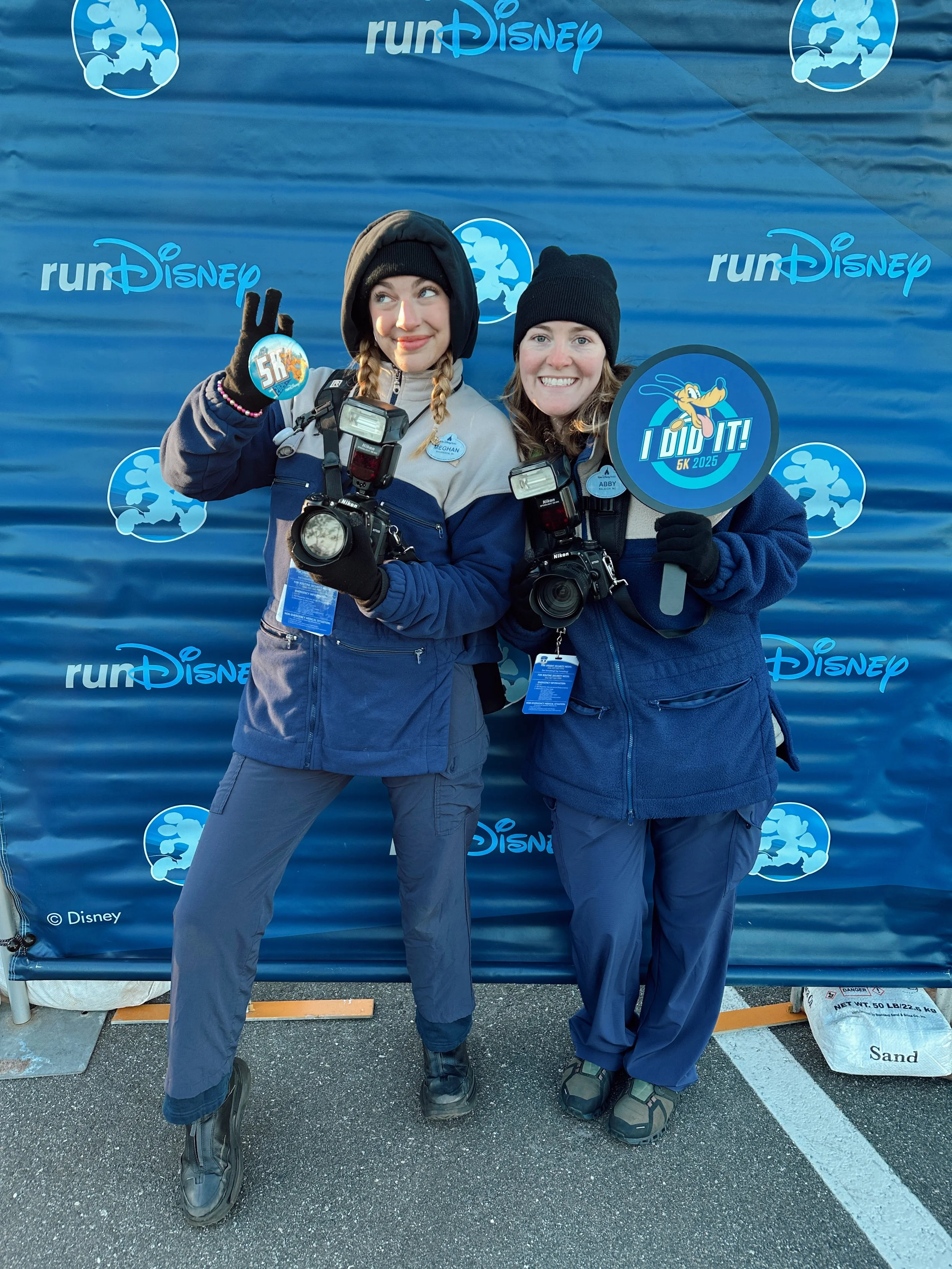 Two women dressed in running gear, smiling, holding medals and cameras, standing in front of a blue Disney-themed backdrop at a running event.