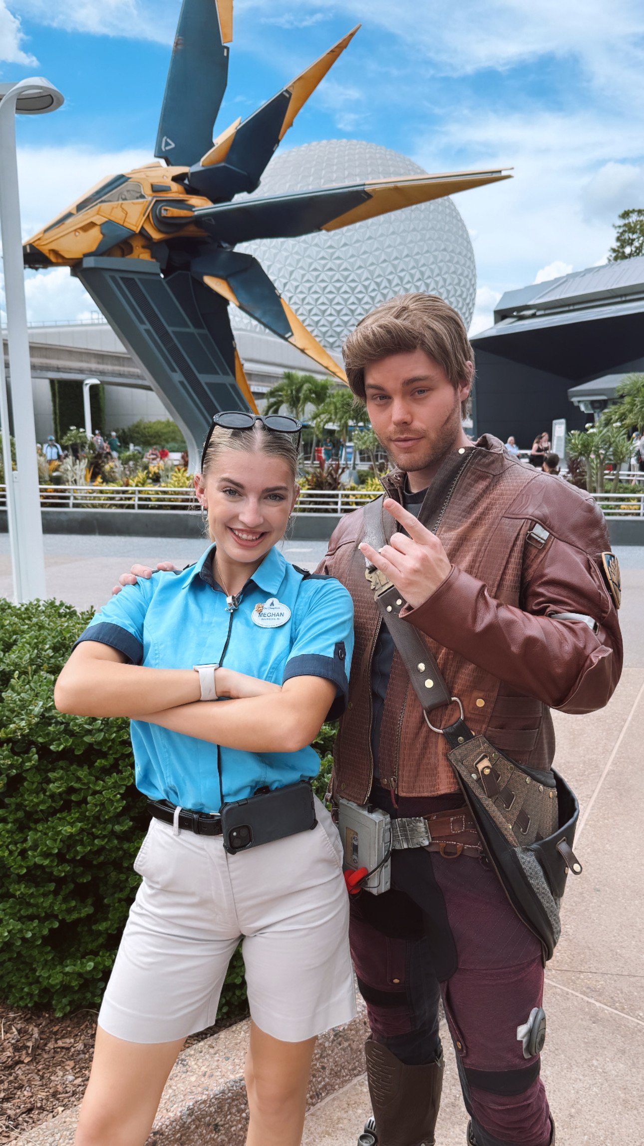 Two people standing together outdoors at a theme park with a large spaceship and a geodesic sphere in the background. The woman on the left is smiling, wearing a blue shirt with a name tag, white shorts, and sunglasses on her head. The man on the right is making a hand gesture, wearing a brown jacket, and has various equipment attached to his belt.