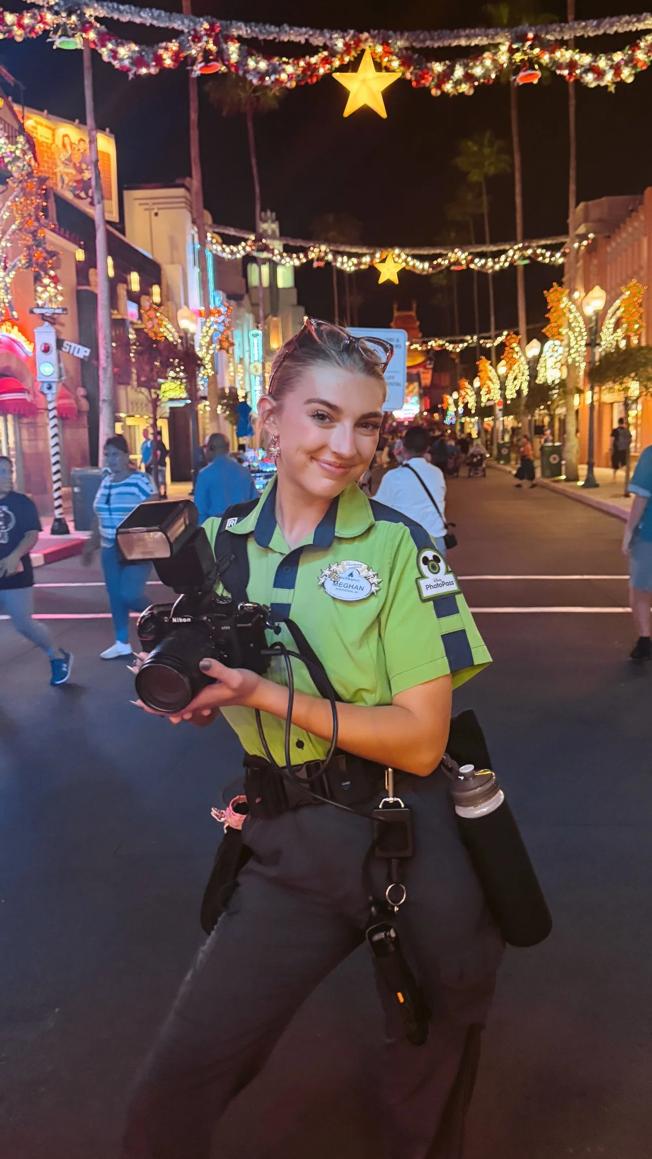 A female police officer in a green uniform holding a camera and smiling at the camera in a festive, decorated street at night with colorful lights, stars, and people walking around.