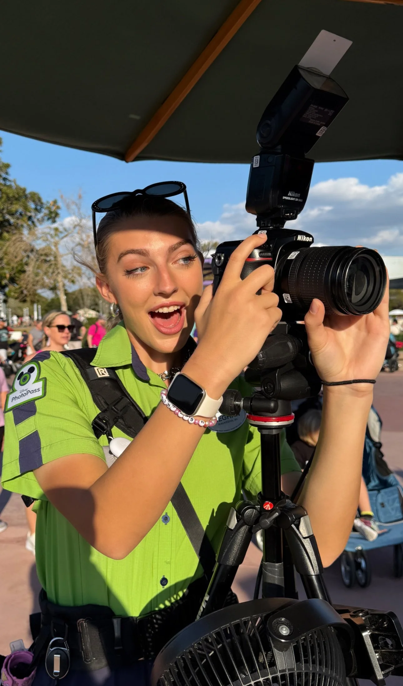 A young woman in a lime green shirt and sunglasses on her head is smiling and looking through a camera mounted on a tripod, outdoors on a sunny day with people and trees in the background.