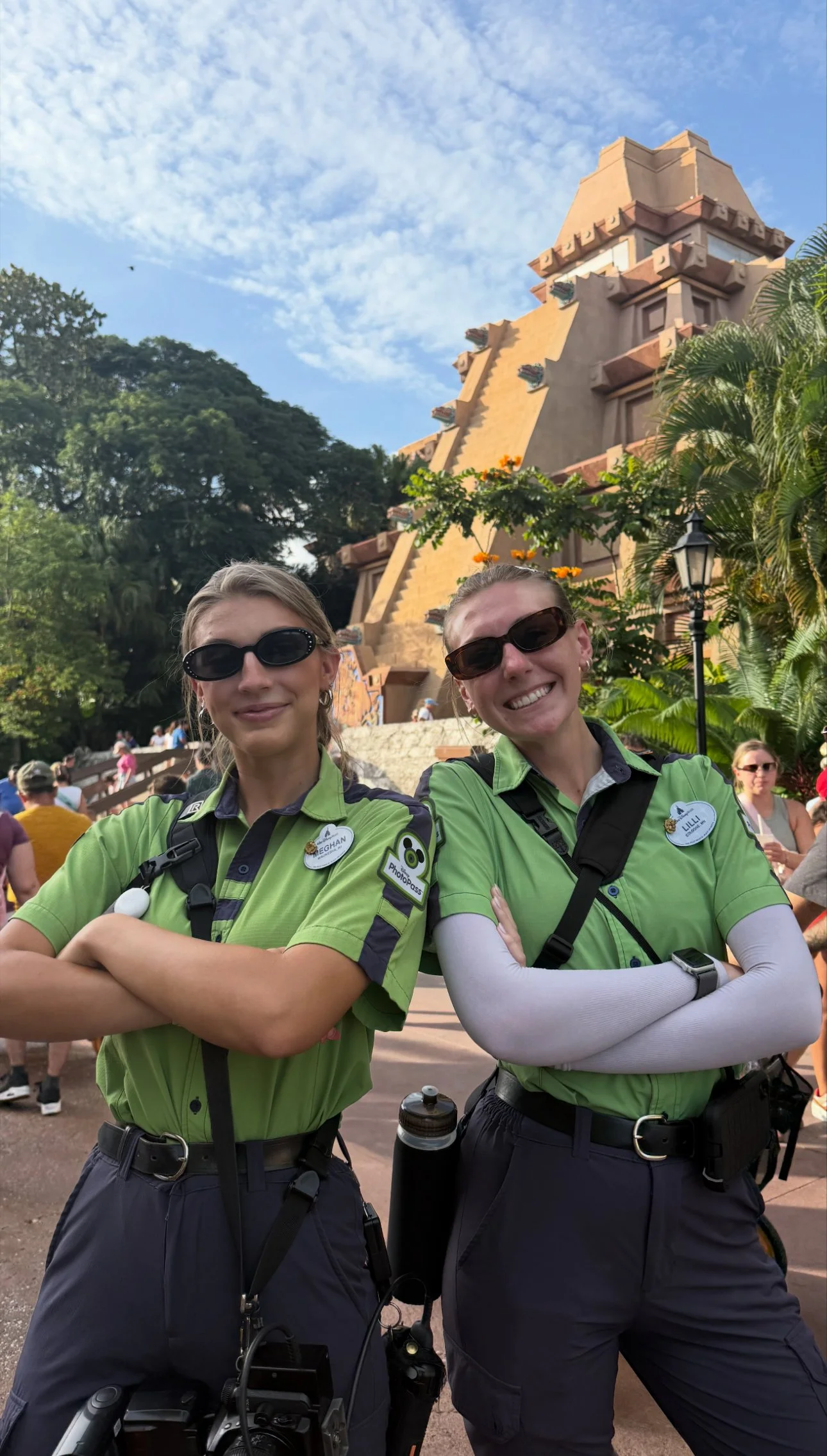 Two women in green security uniform shirts pose with their arms crossed in front of a Mayan pyramid at a theme park.