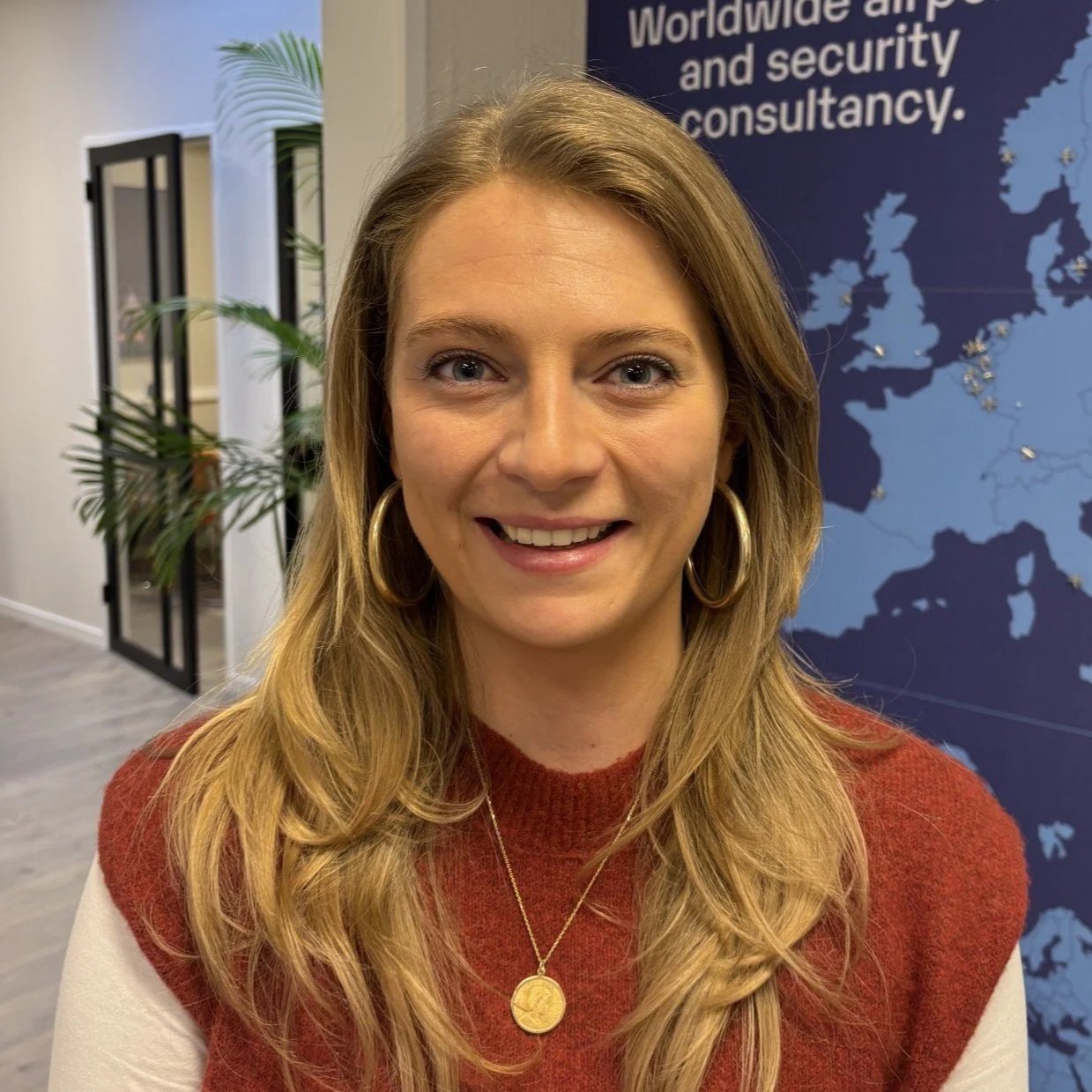 A woman with long wavy blonde hair, wearing a red and white sweater, gold hoop earrings, and a gold necklace with a round pendant, smiling in an office environment with a plant and a world map in the background.