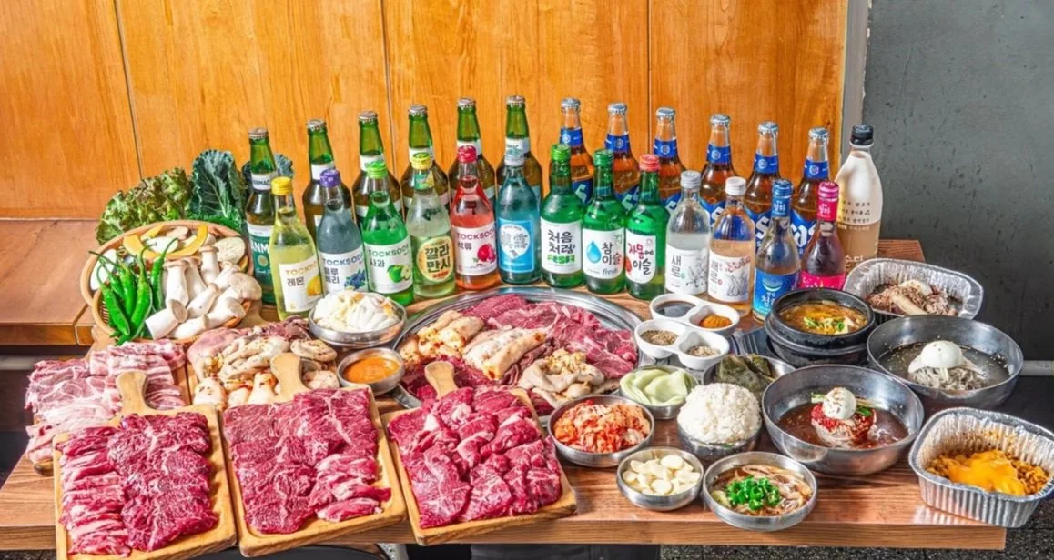 An assortment of Korean dishes including sliced meats, vegetables, sauces, rice, and various bottles of traditional Korean alcoholic beverages at Myeongdong Korean BBQ Mongvely.