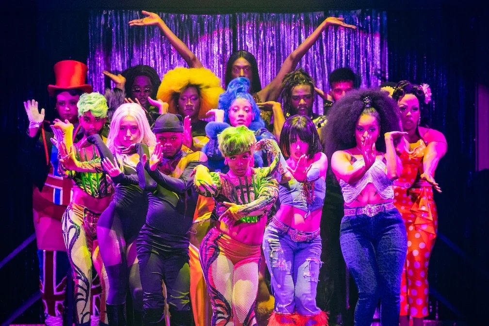 A diverse group of performers in colorful costumes on stage with a purple curtain backdrop, voguing and striking various poses.