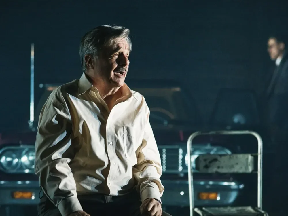 An older man with gray hair and a mustache, sitting and looking distressed, in front of a dark vintage car, with another person blurred in the background.