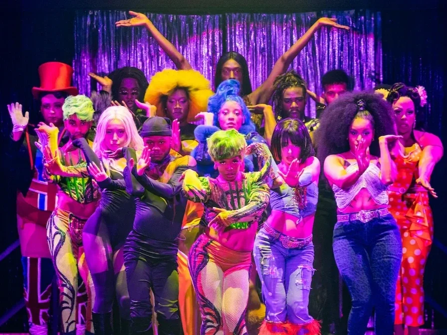 Group of performers in colorful costumes striking dramatic poses on stage with purple curtains in the background.