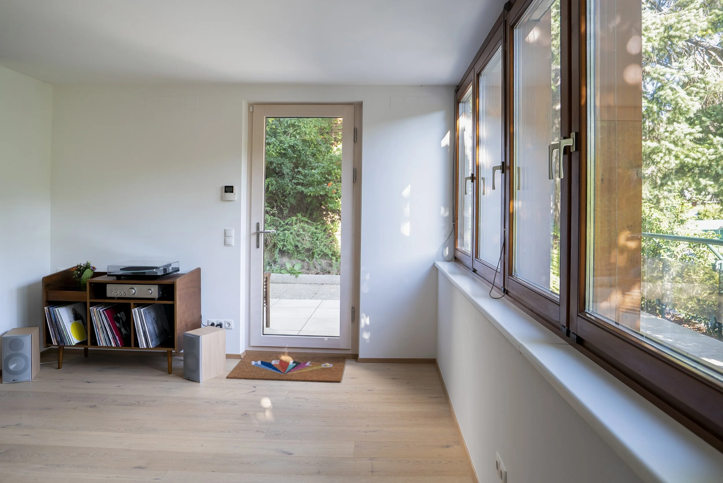 Yogastudio Krottenbachstraße 1190 Wien: Loftartiges Yogastudio mit großen Fenstern, Holzboden, Terrassentüre nach draußen, Plattenspieler auf einem Regal, Schallplatten, Lautsprecher, Willkommensmatte.