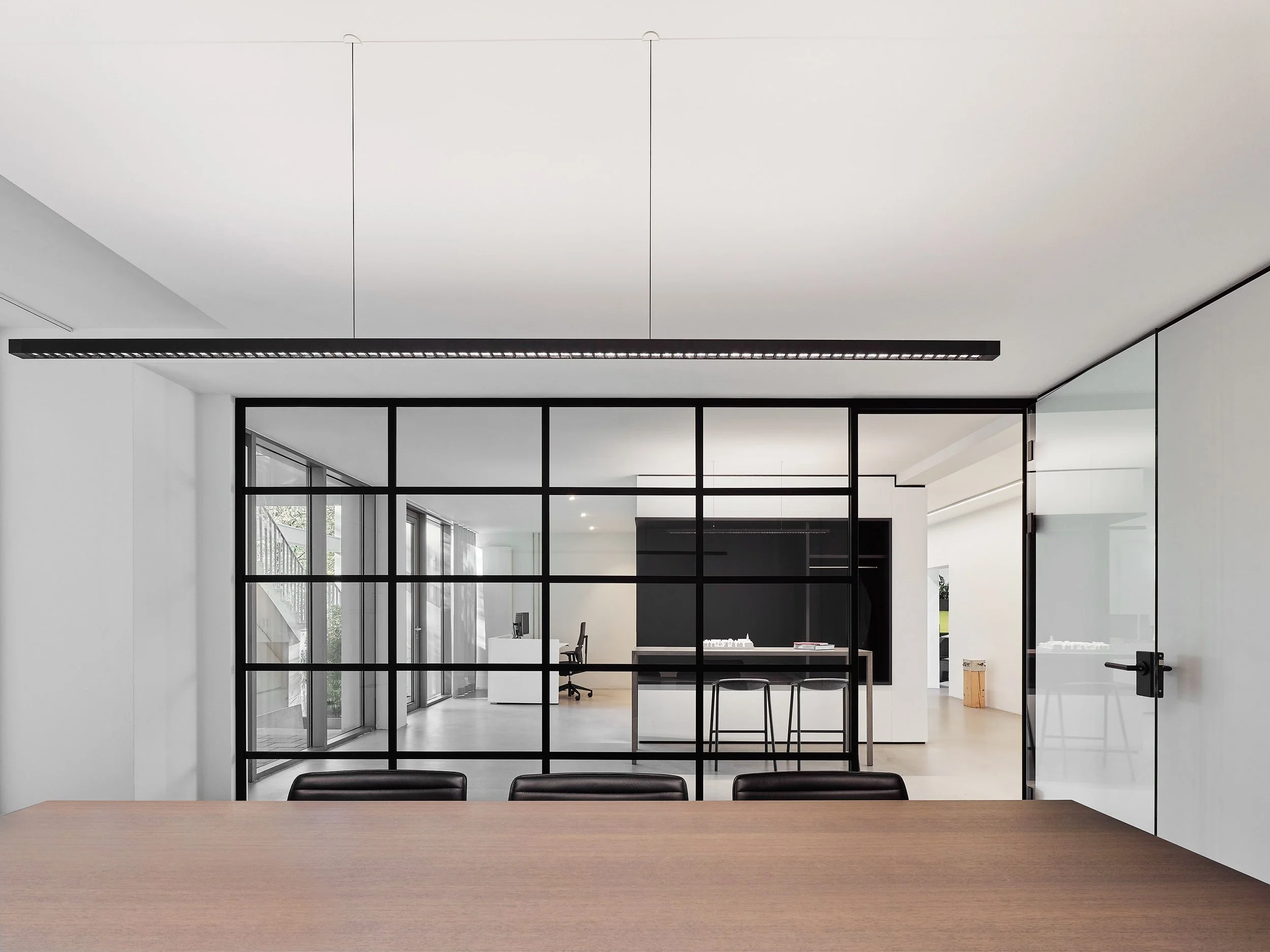 Moderne, minimalistische Bürogestaltung mit schwarzen Metallrahmen, Glaswänden, weißen Wänden und einer Holz-Tischplatte.