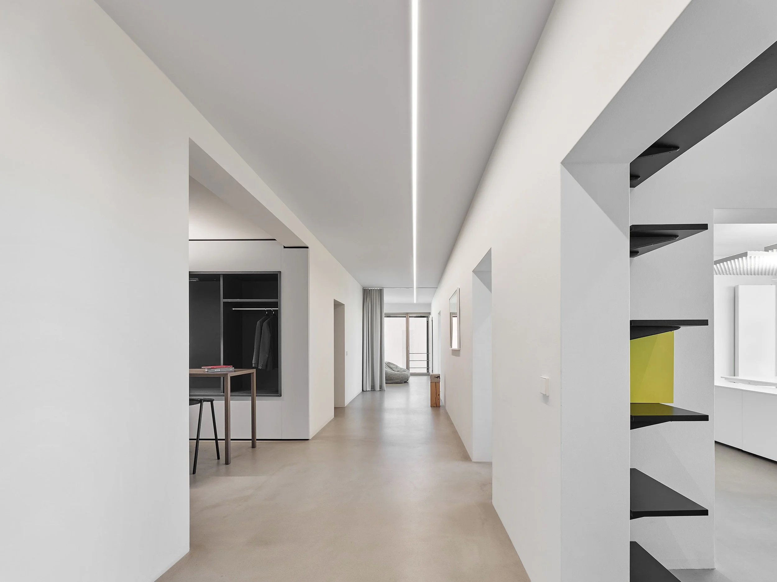 Moderner, minimalistischer Innenraum mit weißen Wänden, Decke und Boden, einem offenen Durchgang, einer Sitzecke bei großen Fenstern, und einer schwarzen Regaleinheit mit bunten Akzenten.