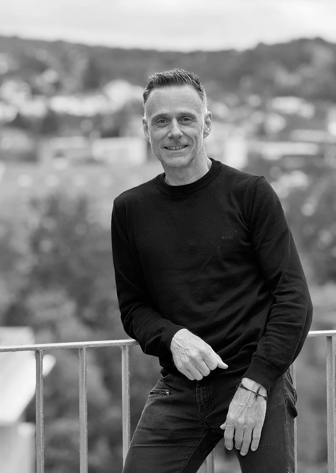 Ein lächelnder Mann in einem schwarzen Pullover steht an einem Geländer, im Hintergrund eine Stadtlandschaft mit Hügeln, alles in Schwarz-Weiß.